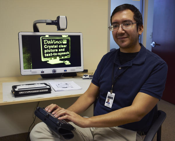 Jose is seated by a computer screen demonstrating enlarged text while he poses his fingers over a Refresher Braille in his lap.
